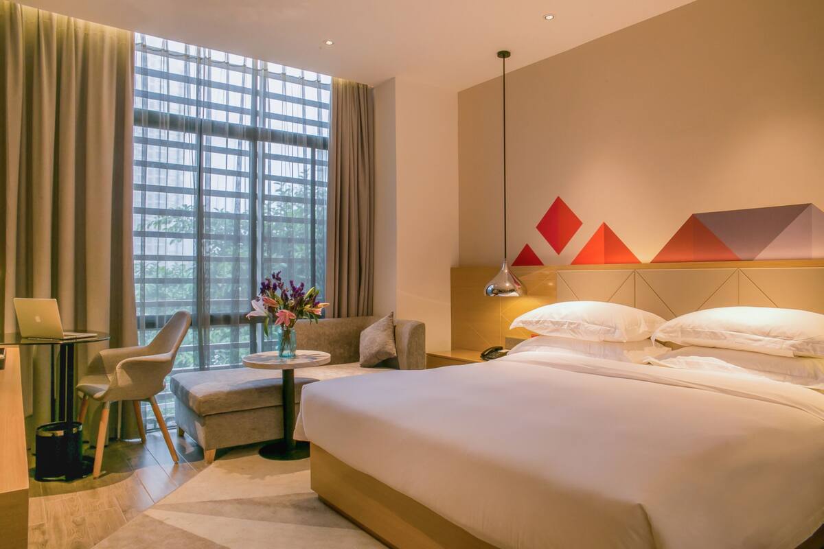 A modern hotel room with a large bed, a desk with a laptop, a sofa, and a view of greenery through the window.