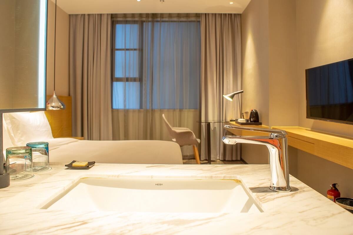 A modern hotel bathroom with a large white bathtub, a sink, and a mirror. There is a window with curtains, a chair, and a television mounted on the wall.