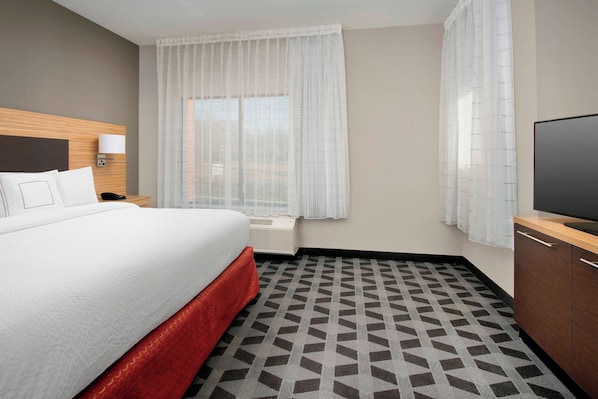 Habitación de hotel con cama, televisor, mesita de noche con lámpara, ventana con cortinas translúcidas y alfombra estampada.