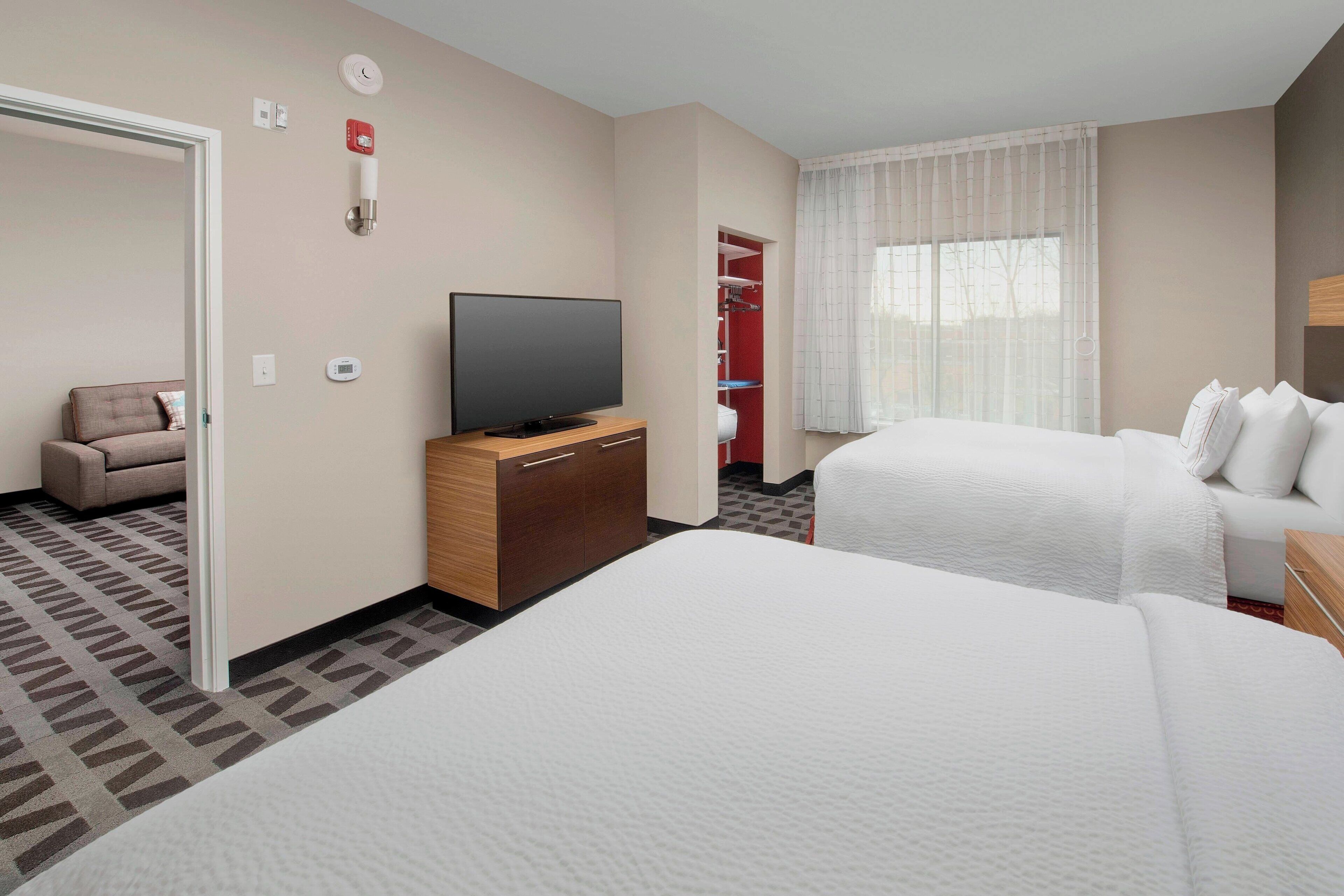 Habitación de hotel con dos camas, un televisor sobre un soporte de madera, un armario rojo y una ventana con cortinas traslúcidas.