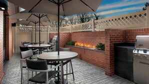 An outdoor patio with a brick barbecue grill, round tables, and chairs, surrounded by a white lattice fence. - TownePlace Suites by Marriott College Park (College Park)
