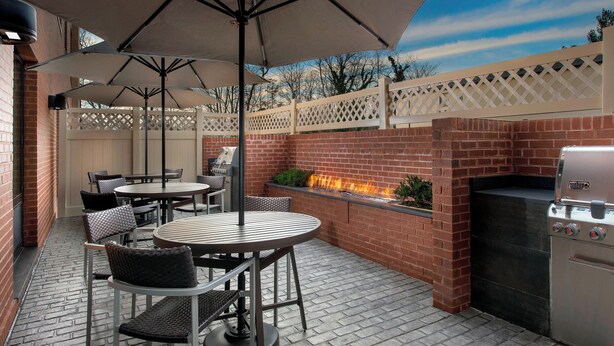 An outdoor patio with a brick barbecue grill, round tables, and chairs, surrounded by a white lattice fence. - TownePlace Suites by Marriott College Park (College Park)