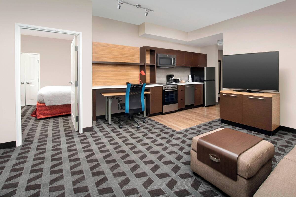 A modern hotel room with a kitchenette, a flat-screen TV, a sofa, and a patterned carpet.