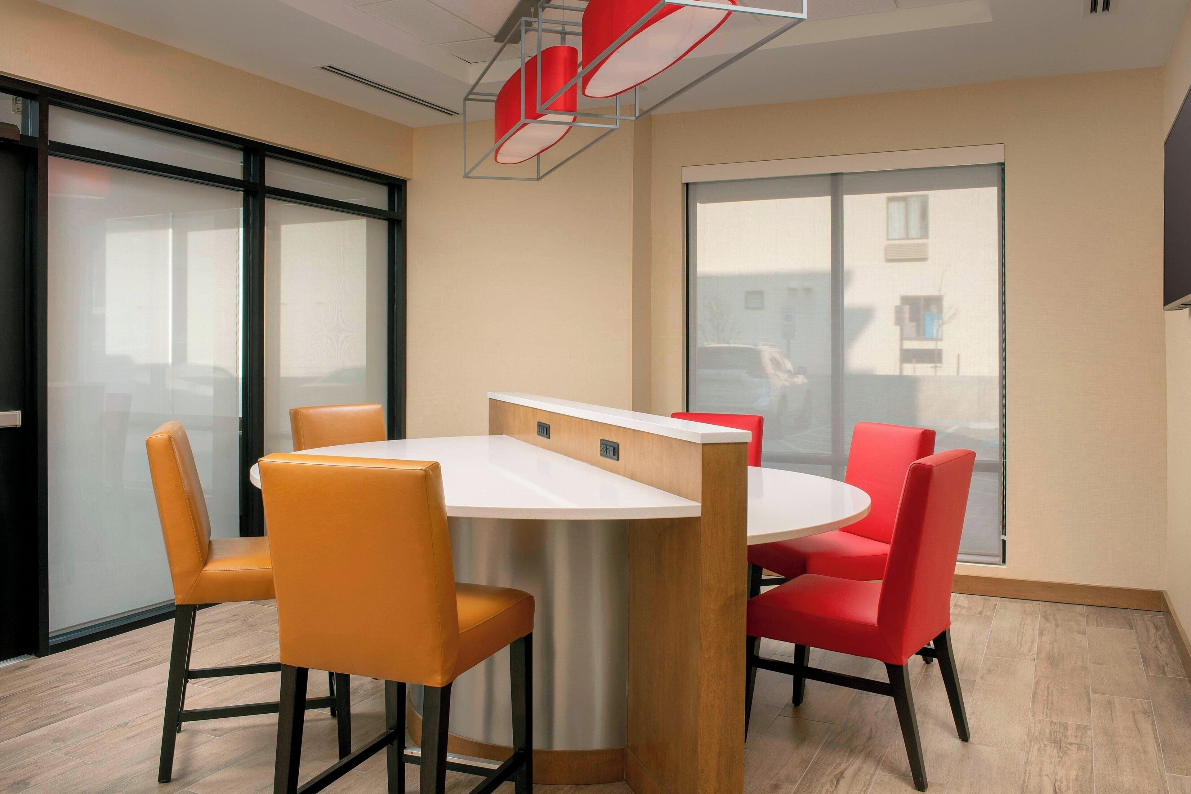 A modern meeting room with a round table, orange and red chairs, large windows, and a ceiling light fixture.
