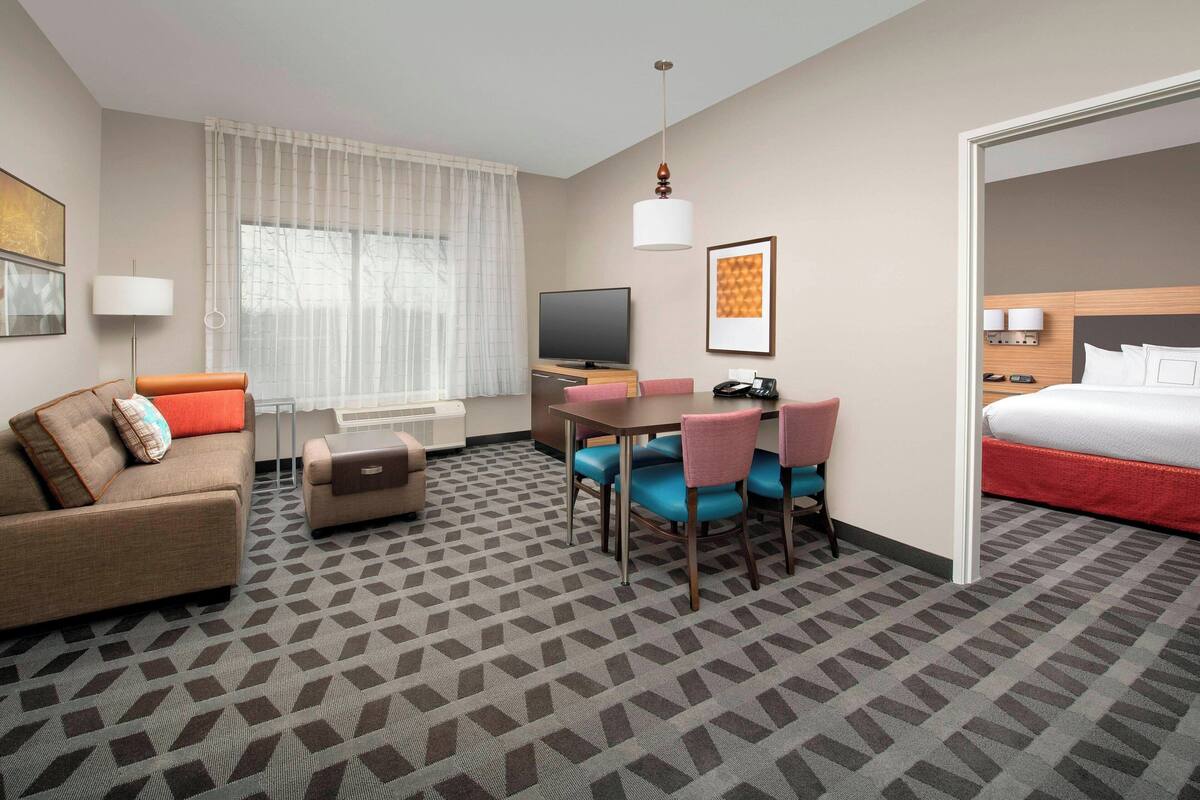 A hotel room with a sofa, a dining table with chairs, a TV, a bed, and a patterned carpet.