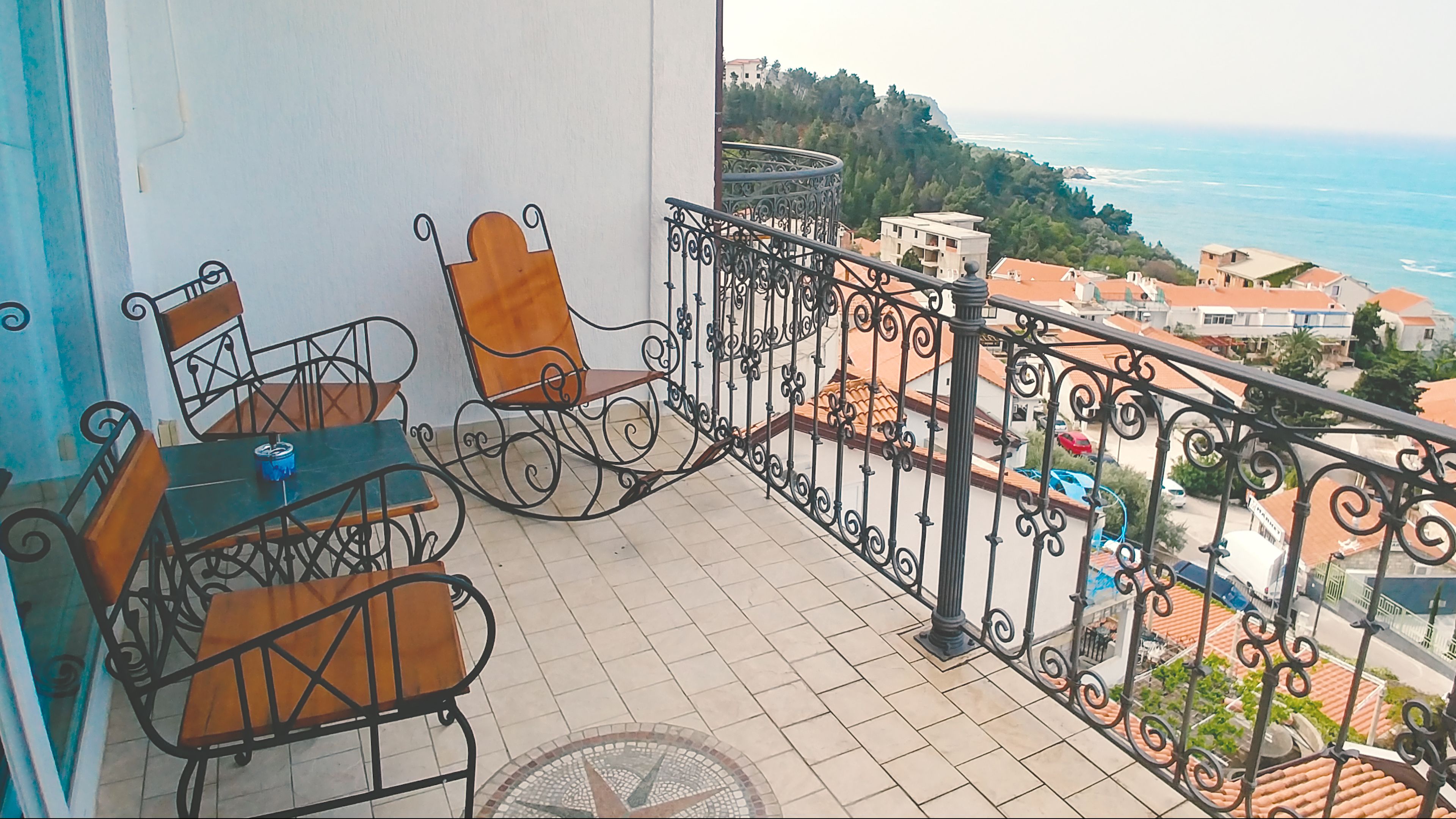A balcony with a table and chairs, a view of the sea, and buildings in the background.