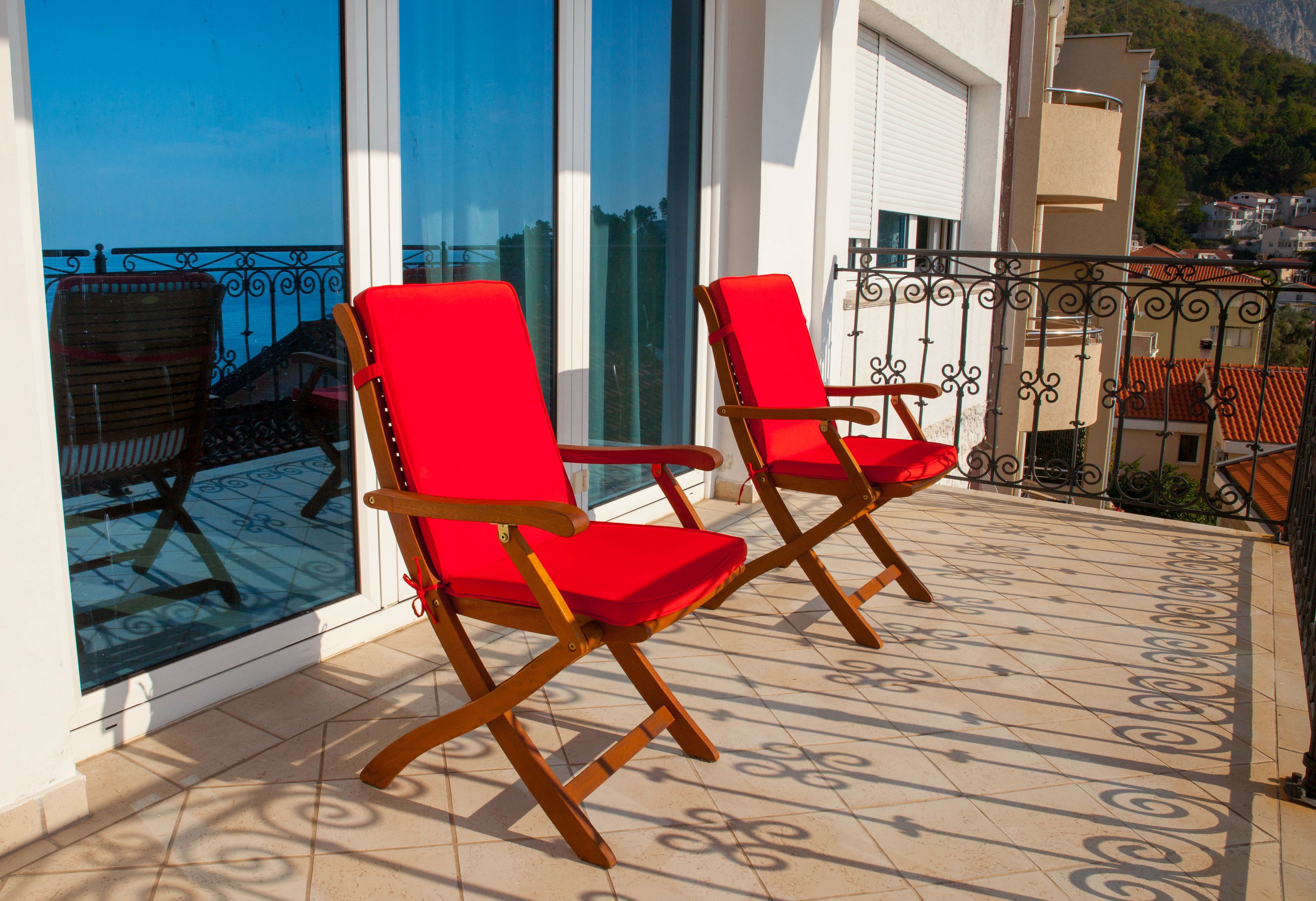 Deux fauteuils rouges sont placés sur un balcon carrelé, d’où l’on peut admirer la mer et, au loin, une montagne.