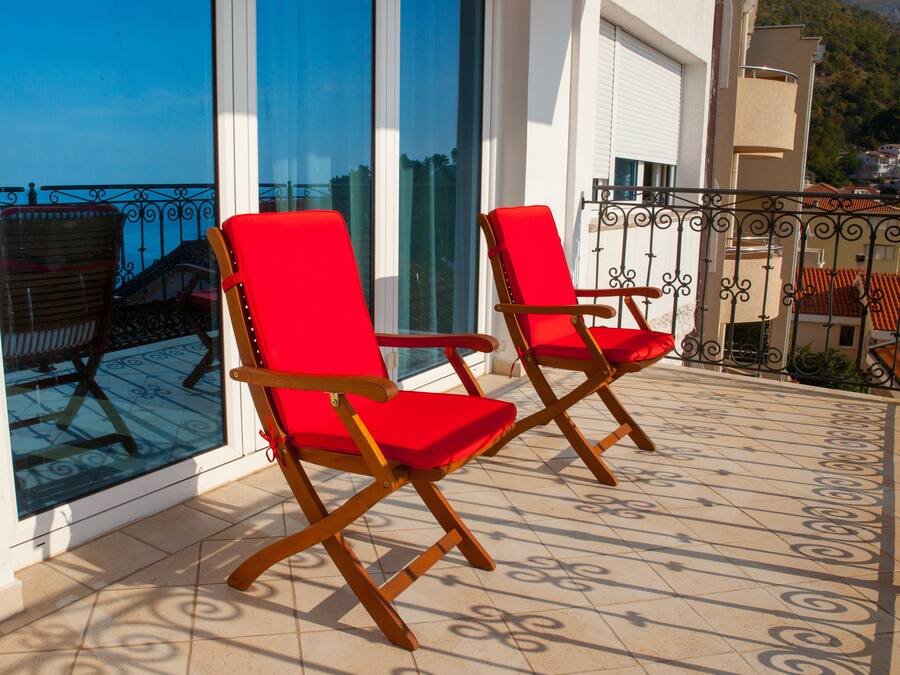 Deux fauteuils rouges sont placés sur un balcon carrelé, d’où l’on peut admirer la mer et, au loin, une montagne.