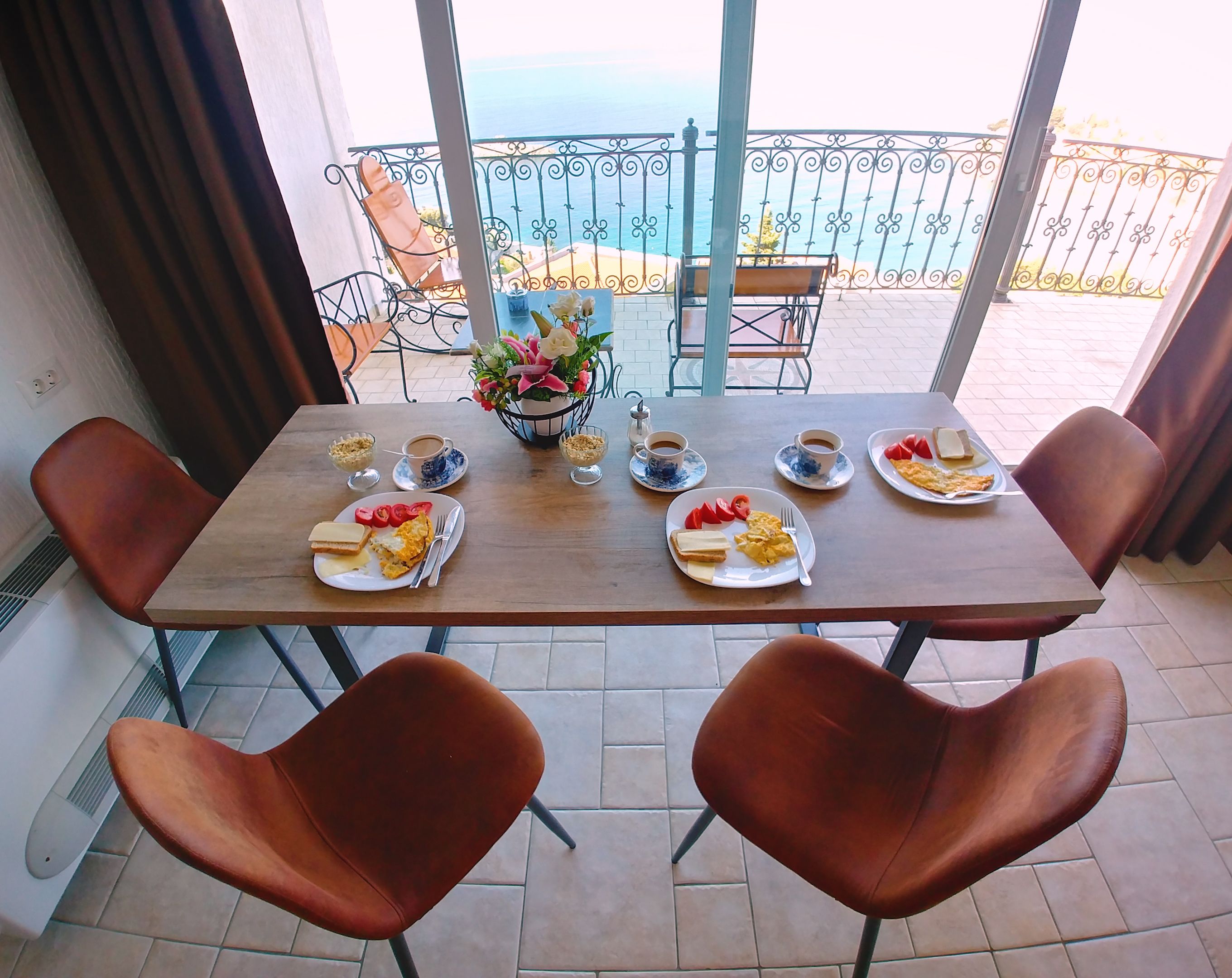 A dining table with four plates of breakfast, two chairs, a flower vase, and a view of the sea.