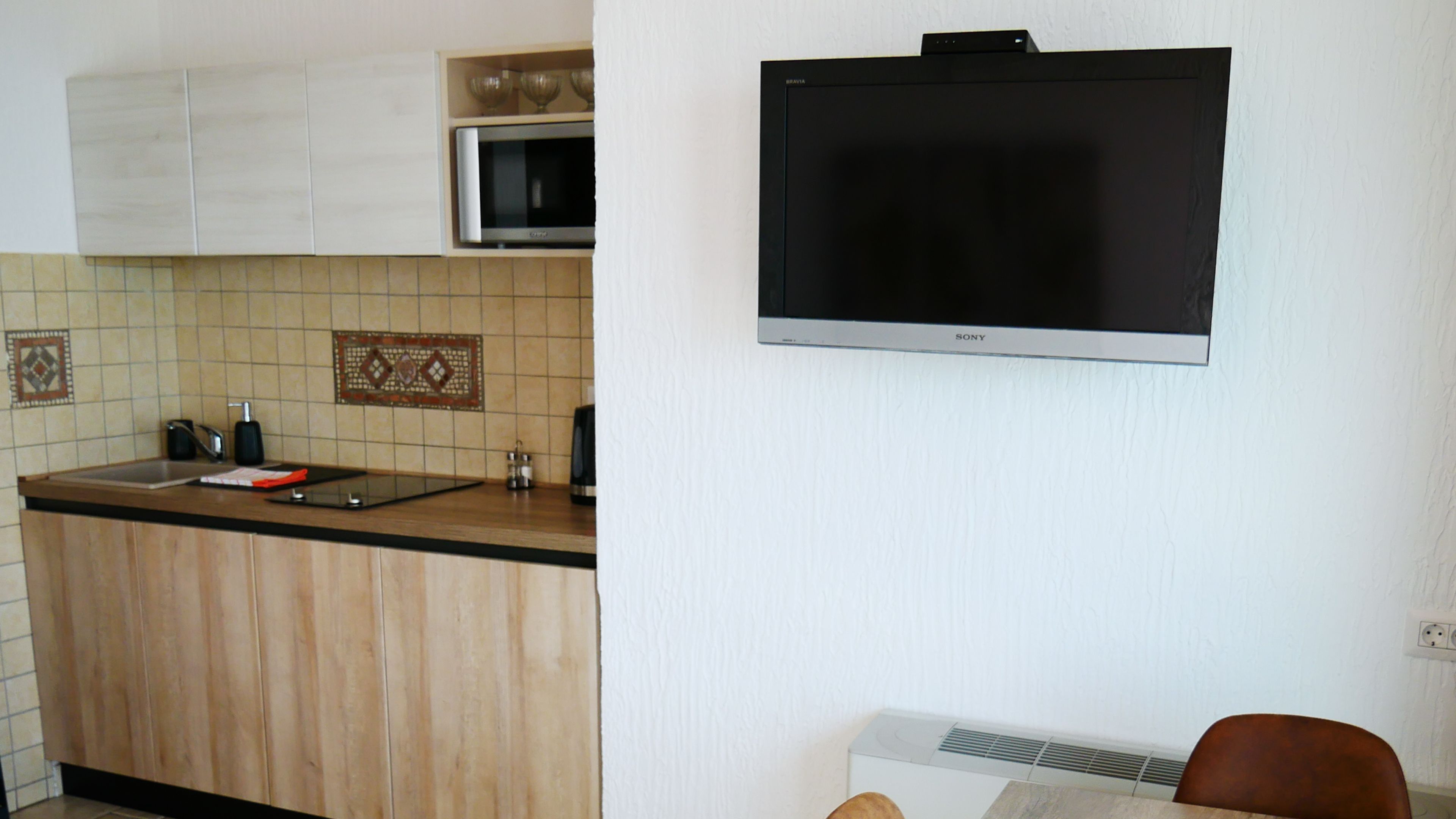 A kitchen with wooden cabinets, a microwave, a flat-screen TV, and a small table with a chair.
