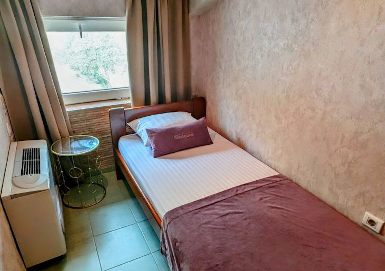 Une petite pièce, de taille modeste, comprenant un lit simple, une table de chevet, une lampe et une fenêtre avec des rideaux.
