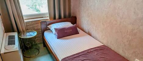 A small, compact room with a single bed, a bedside table, a lamp, and a window with curtains.