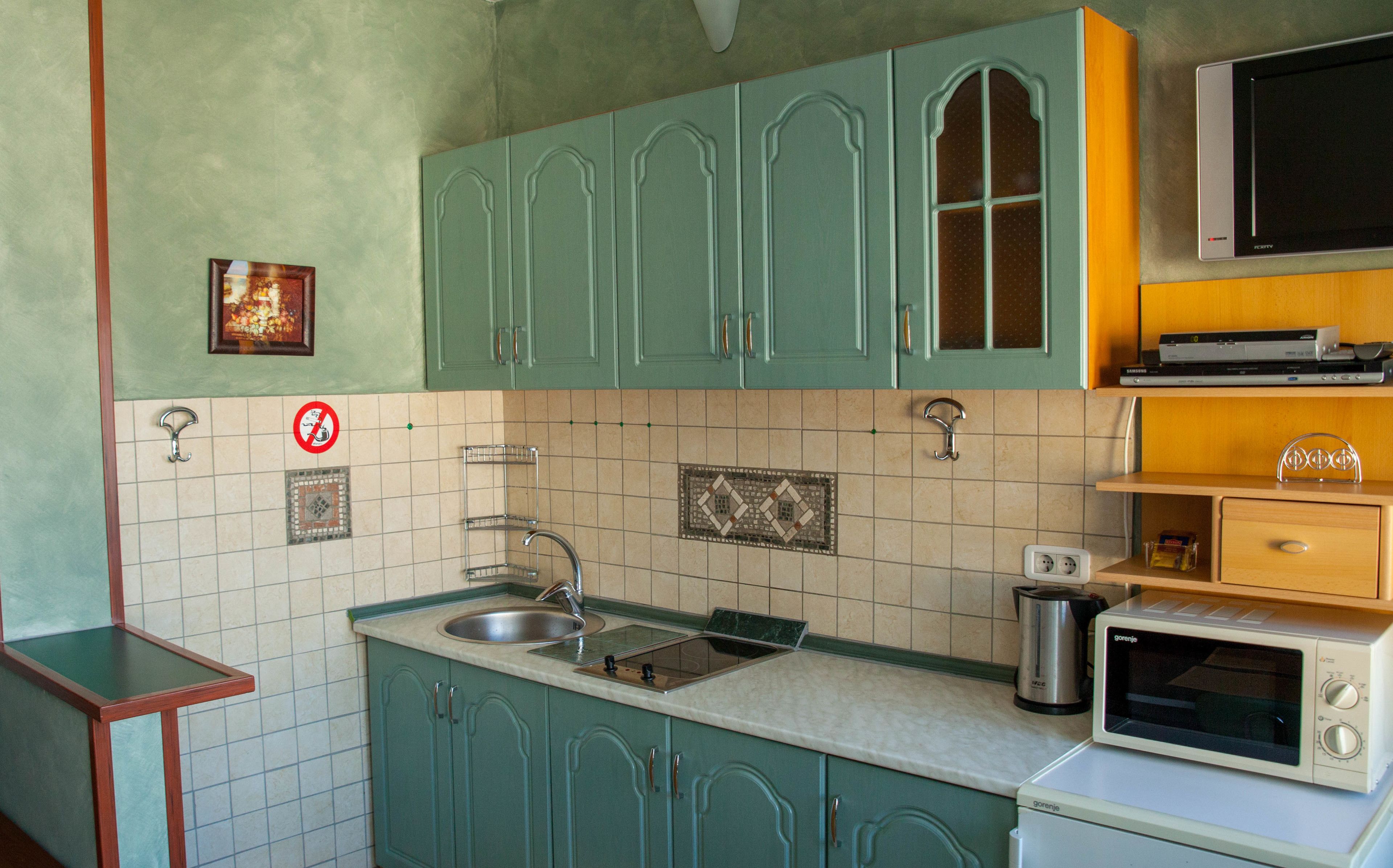 Una cocina con armarios color turquesa, un lavamanos, un microondas y un televisor sobre una estantería de madera.