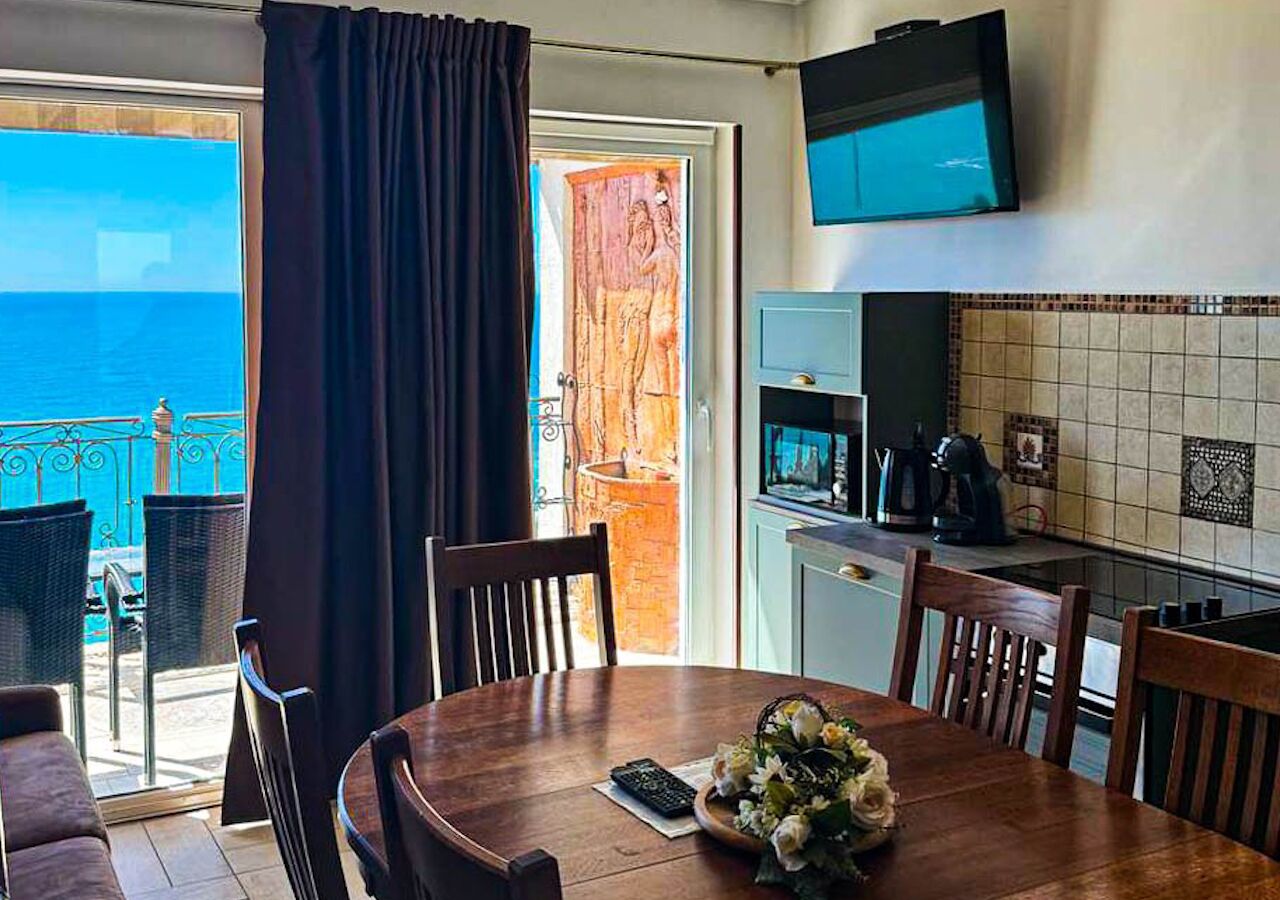 Un coin repas avec une table et des chaises, une cuisine équipée d’un téléviseur et une vue sur la mer par une fenêtre.