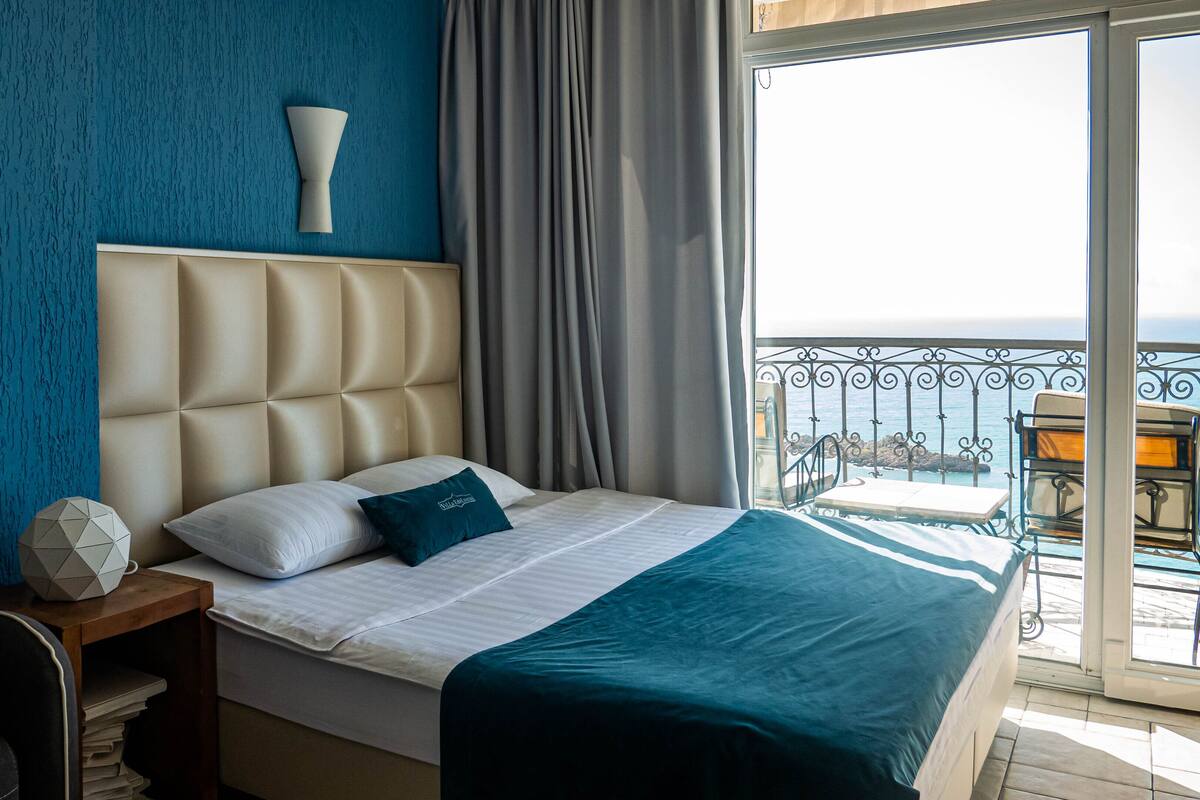 Ein Hotelzimmer mit Bett, Nachttisch, Sessel und Meerblick vom Balkon.