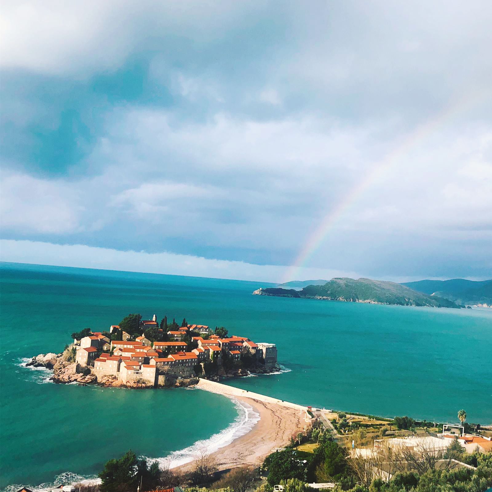 A coastal town with buildings on a peninsula, a beach, and a rainbow in the distance.