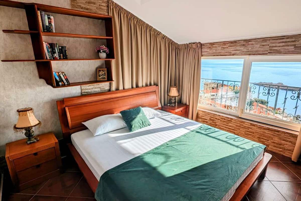 Ein Hotelzimmer mit einem großen Bett, einem Holzkopfteil, einem Nachttisch mit Lampe, einem Bücherregal und Meerblick vom Balkon.