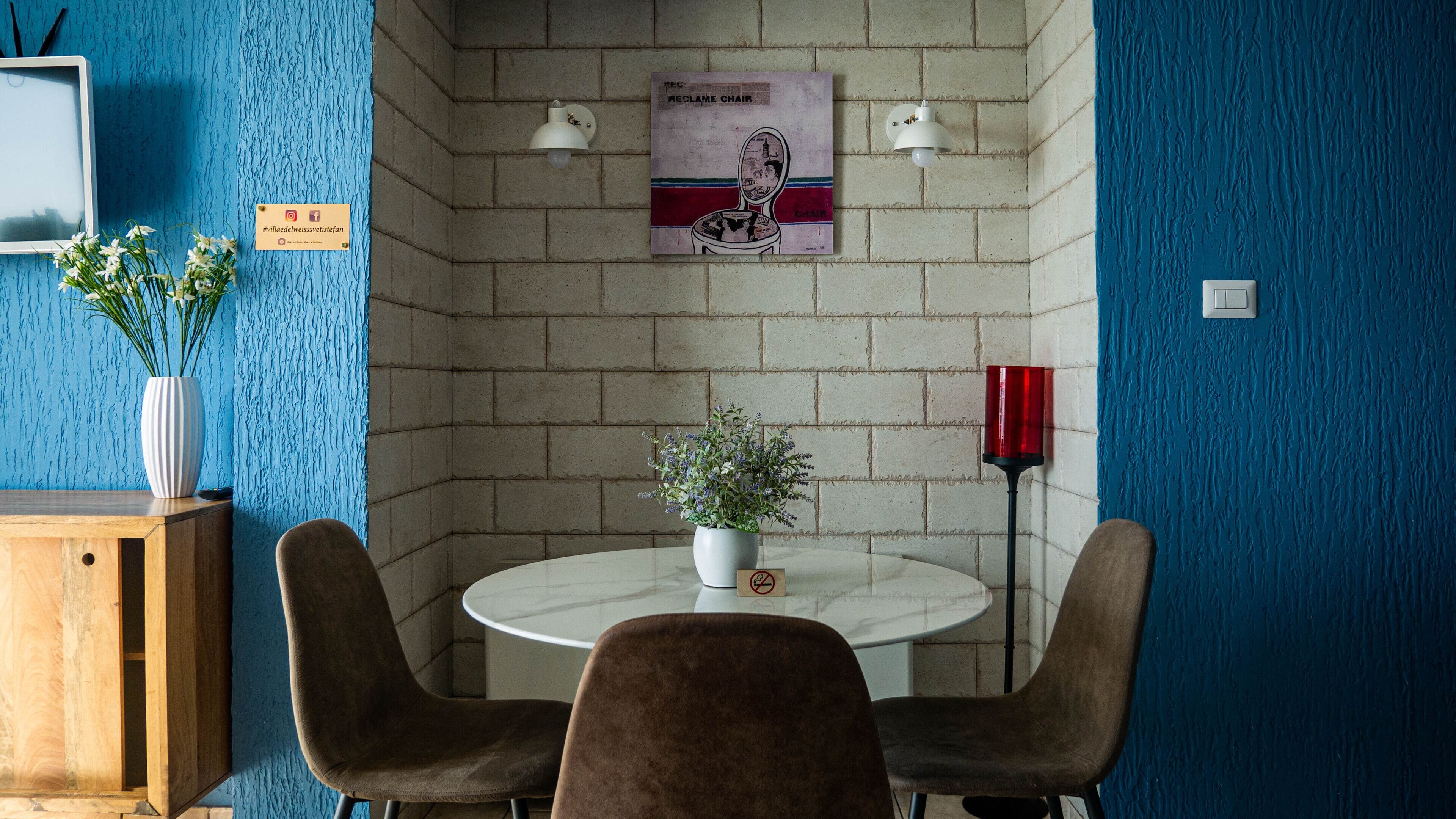 Une pièce avec une table ronde, deux chaises, un vase de fleurs, une affiche au mur et un meuble avec une pancarte.