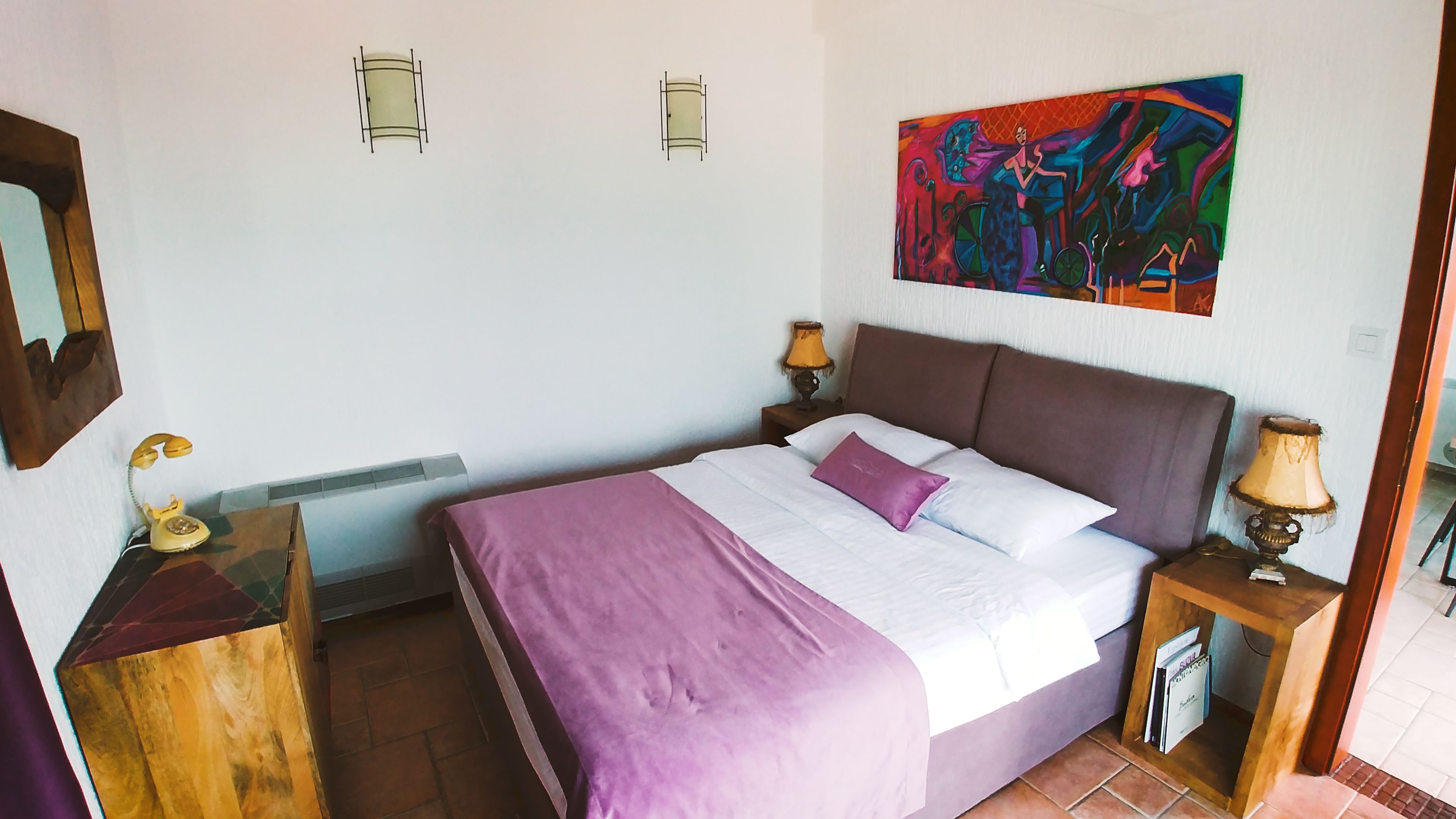 A bedroom with a bed, a nightstand, a lamp, a telephone, and a painting on the wall.