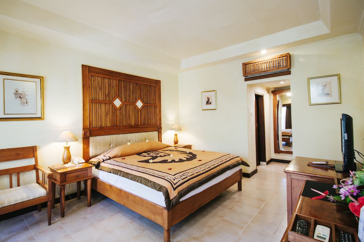 A hotel room with a wooden bed, two bedside tables with lamps, a wooden chair, a desk with a computer, and framed pictures on the walls.