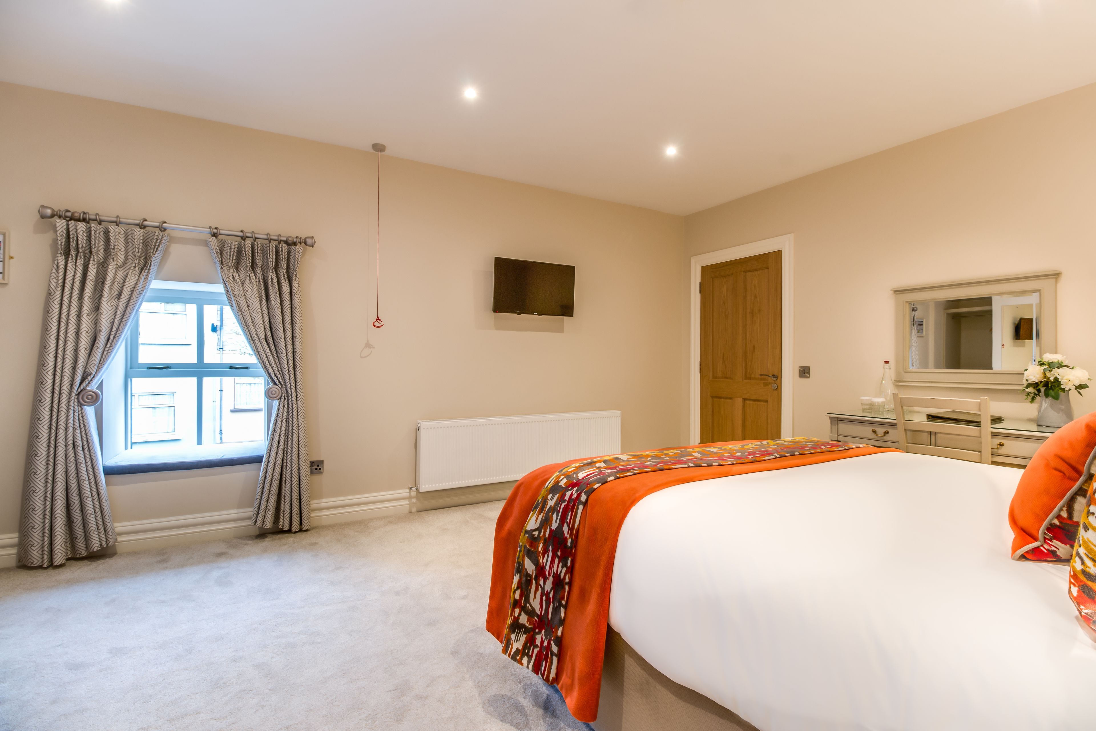 A bedroom with a large bed, a television mounted on the wall, a window with curtains, a radiator, and a door.
