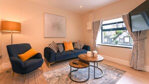 A modern living room with a dark blue armchair, a blue sofa with patterned cushions, a round coffee table, and a large window with curtains. - Kingstons Townhouse (Killorglin)