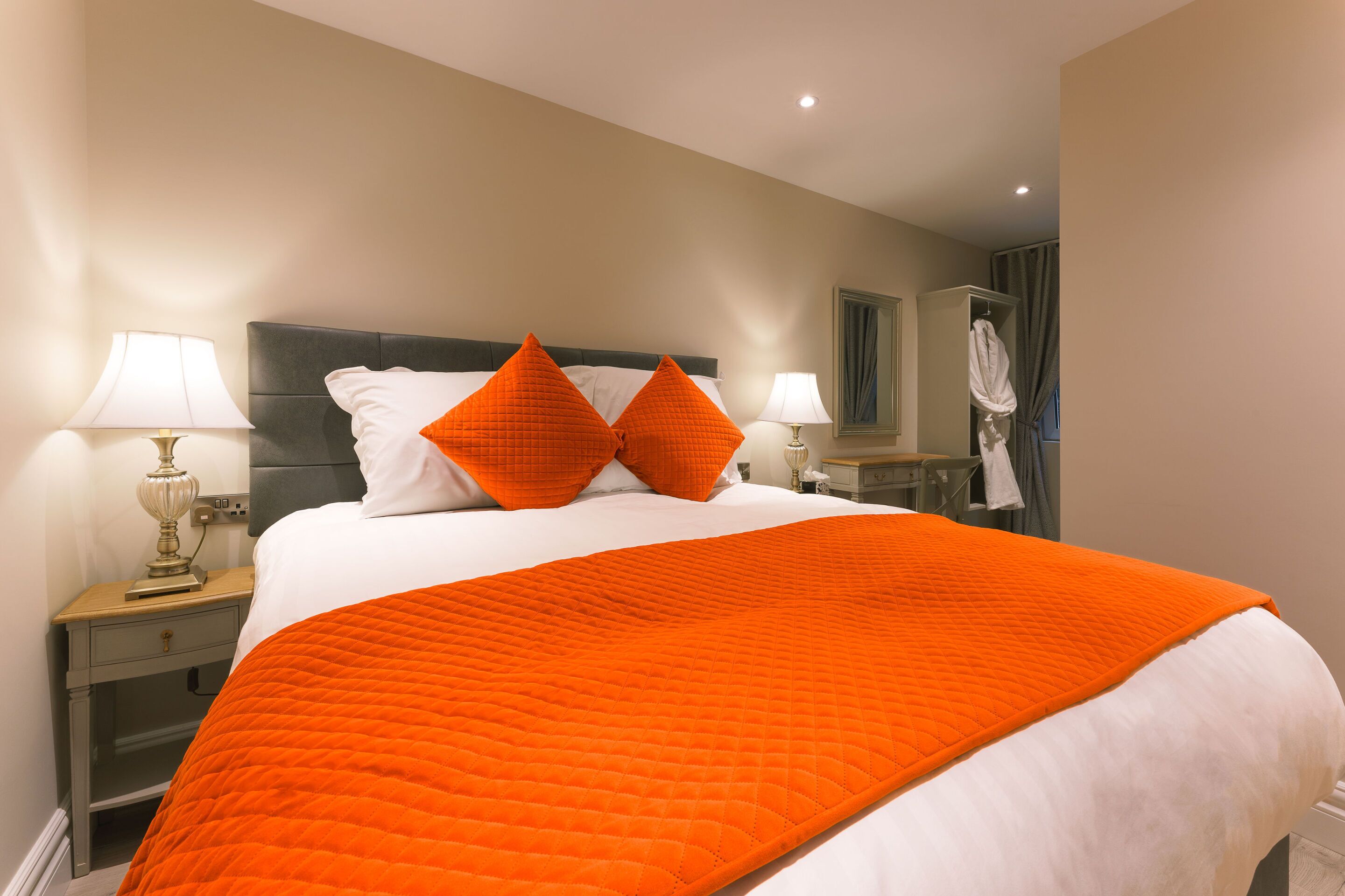 Een netjes opgemaakt bed met een wit-oranje deken, twee witte kussens en twee oranje kussens. Er staat een nachtkastje met een lamp en een klein tafeltje met een spiegel en een badjas.