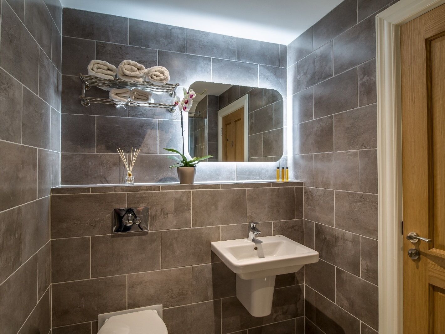 Un baño con baldosas grises, un lavabo, un espejo, un perchero con toallas, una planta en maceta y una pequeña estantería con artículos de aseo.
