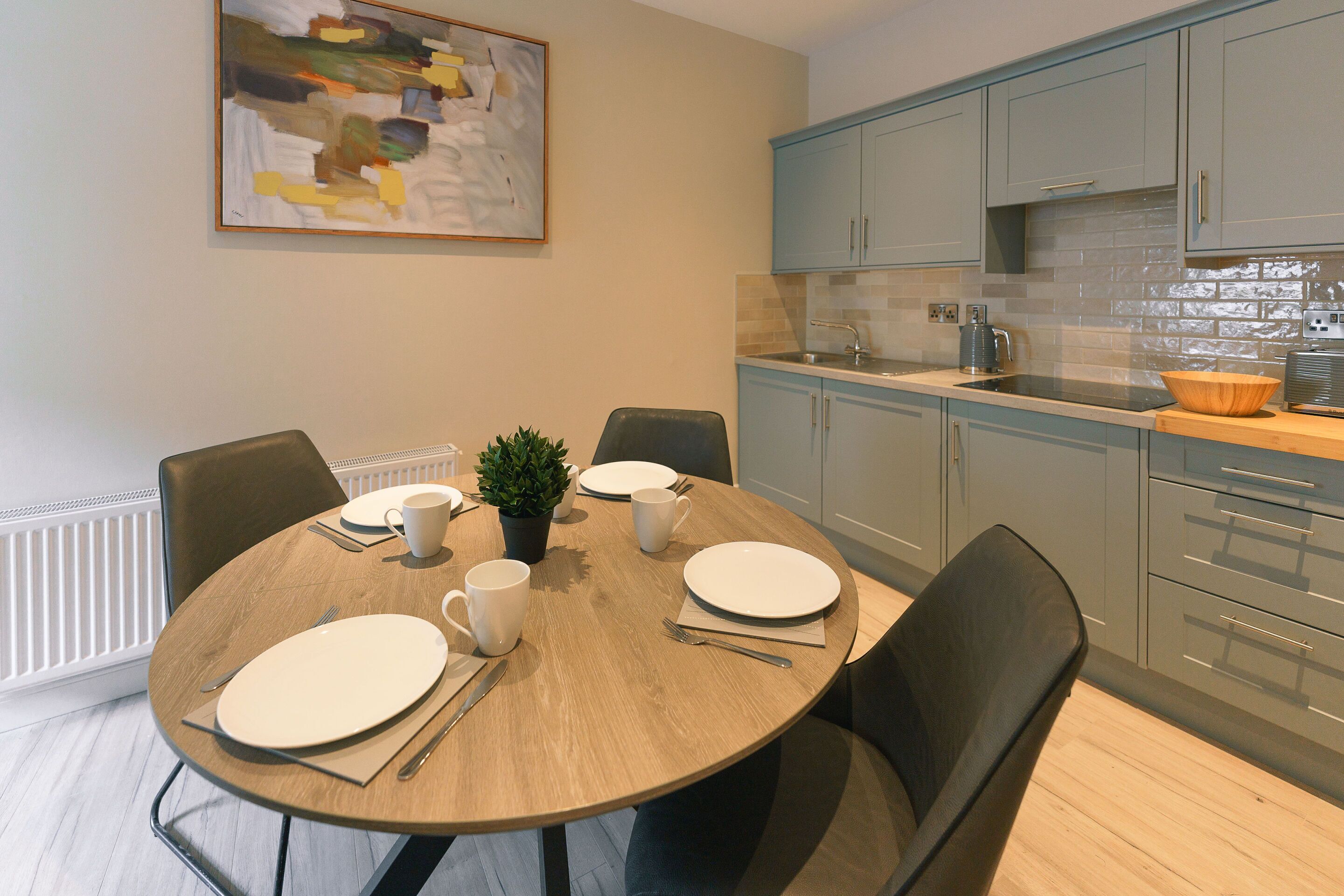 Un comedor con una mesa redonda dispuesta para cuatro personas, rodeada de sillas, una planta en maceta y un cuadro en la pared.