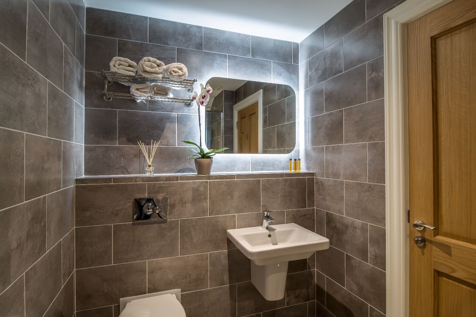 A modern bathroom with dark tiles, a white sink, a mirror, and a wooden door.