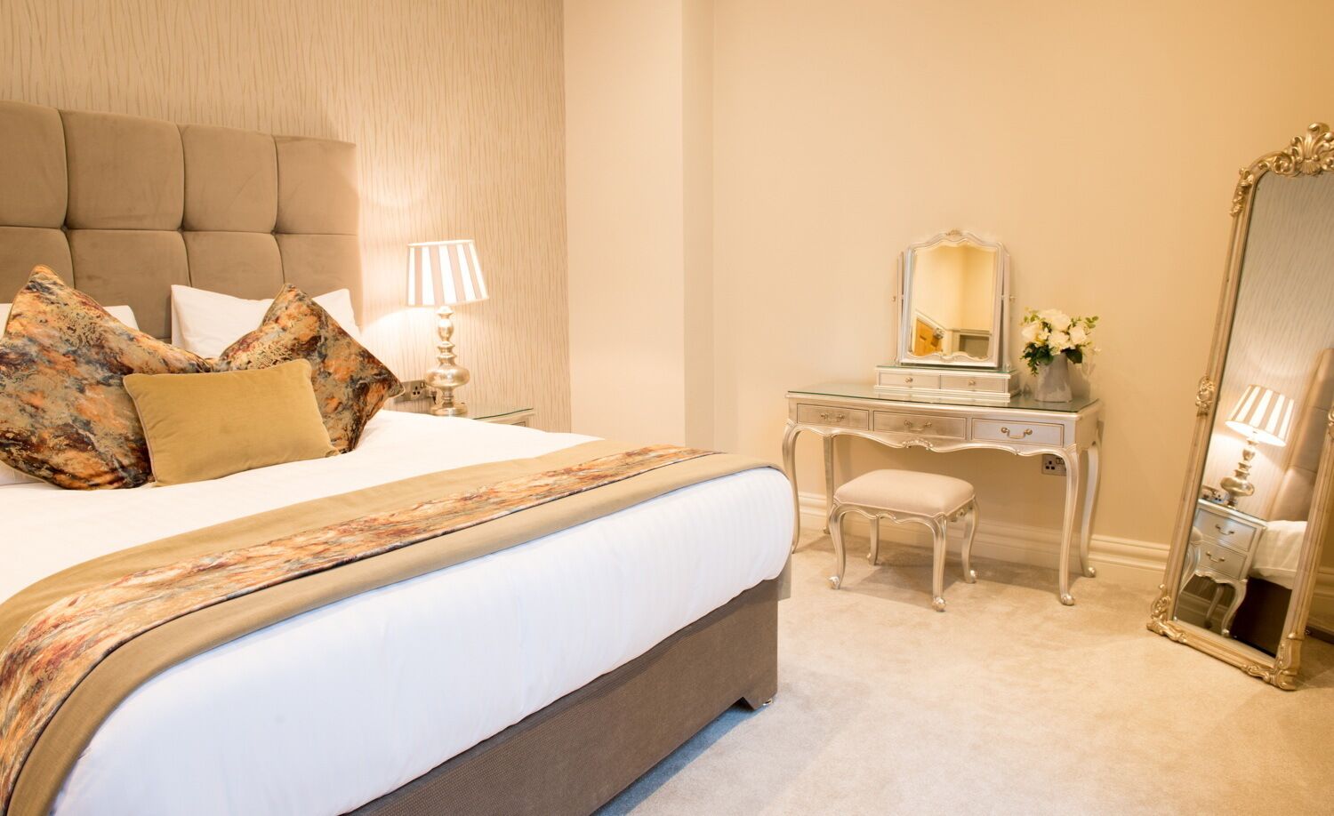 A neatly arranged bedroom with a large bed, a dresser, a mirror, and a lamp.