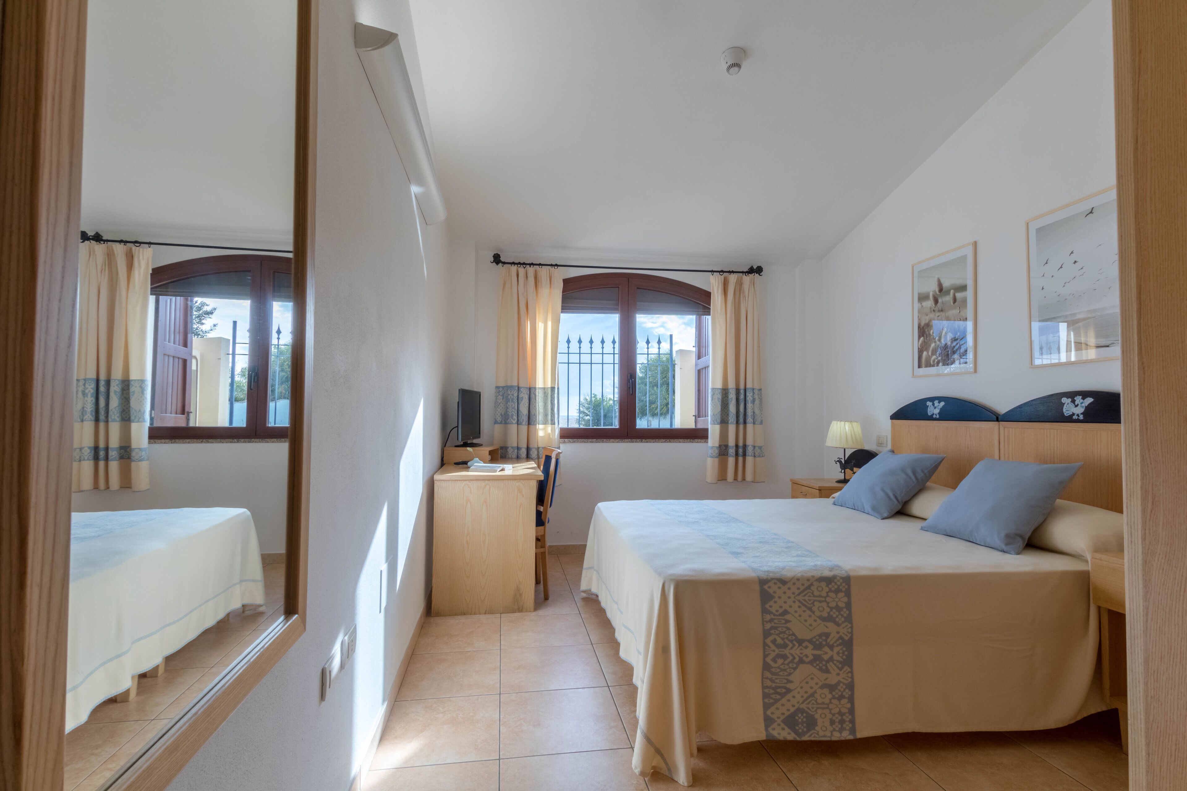 A bedroom with a bed, a desk, and a chair. There are two windows with curtains, a wall-mounted TV, and a framed picture.