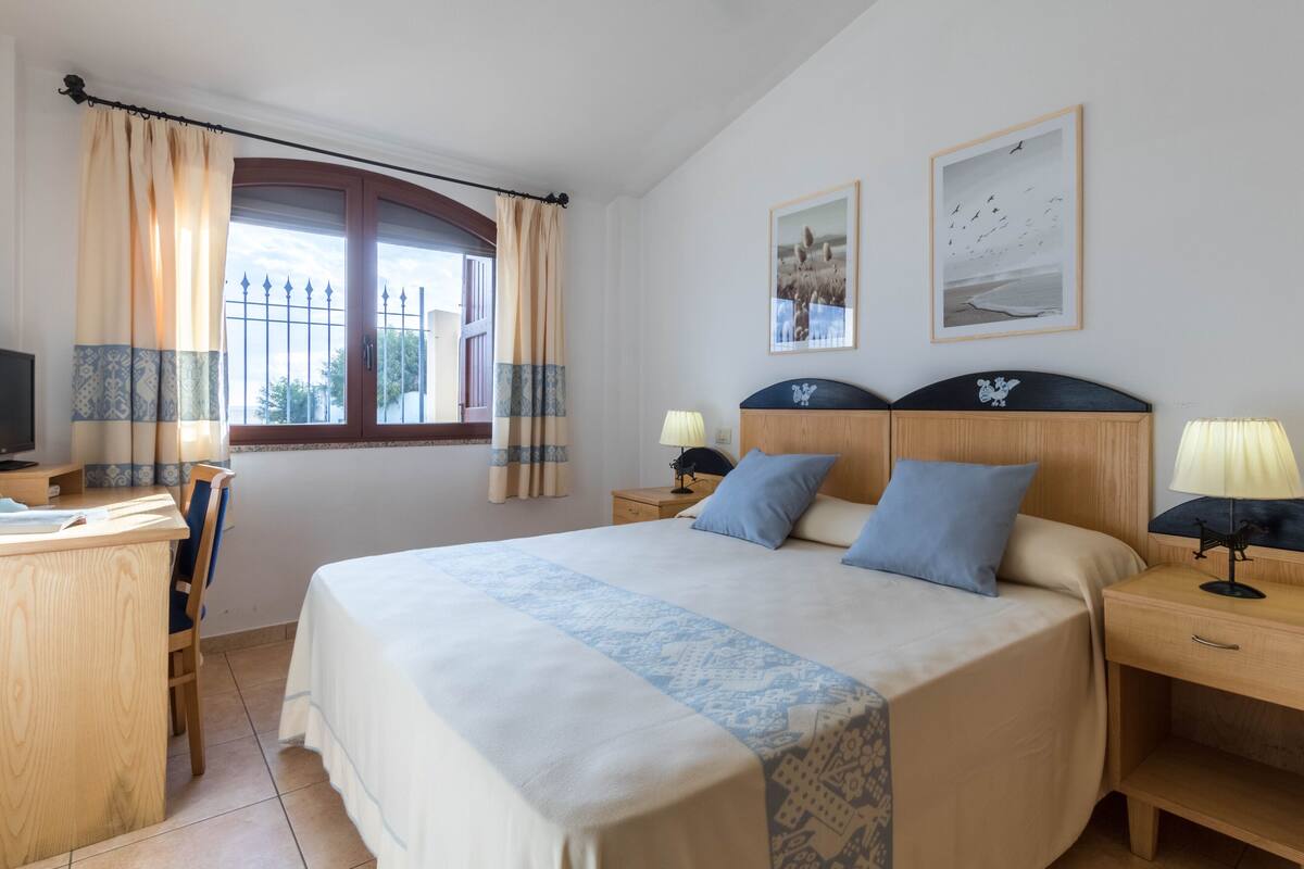 A hotel room with a large bed, two bedside tables with lamps, a desk with a chair, a TV, and a window with curtains.