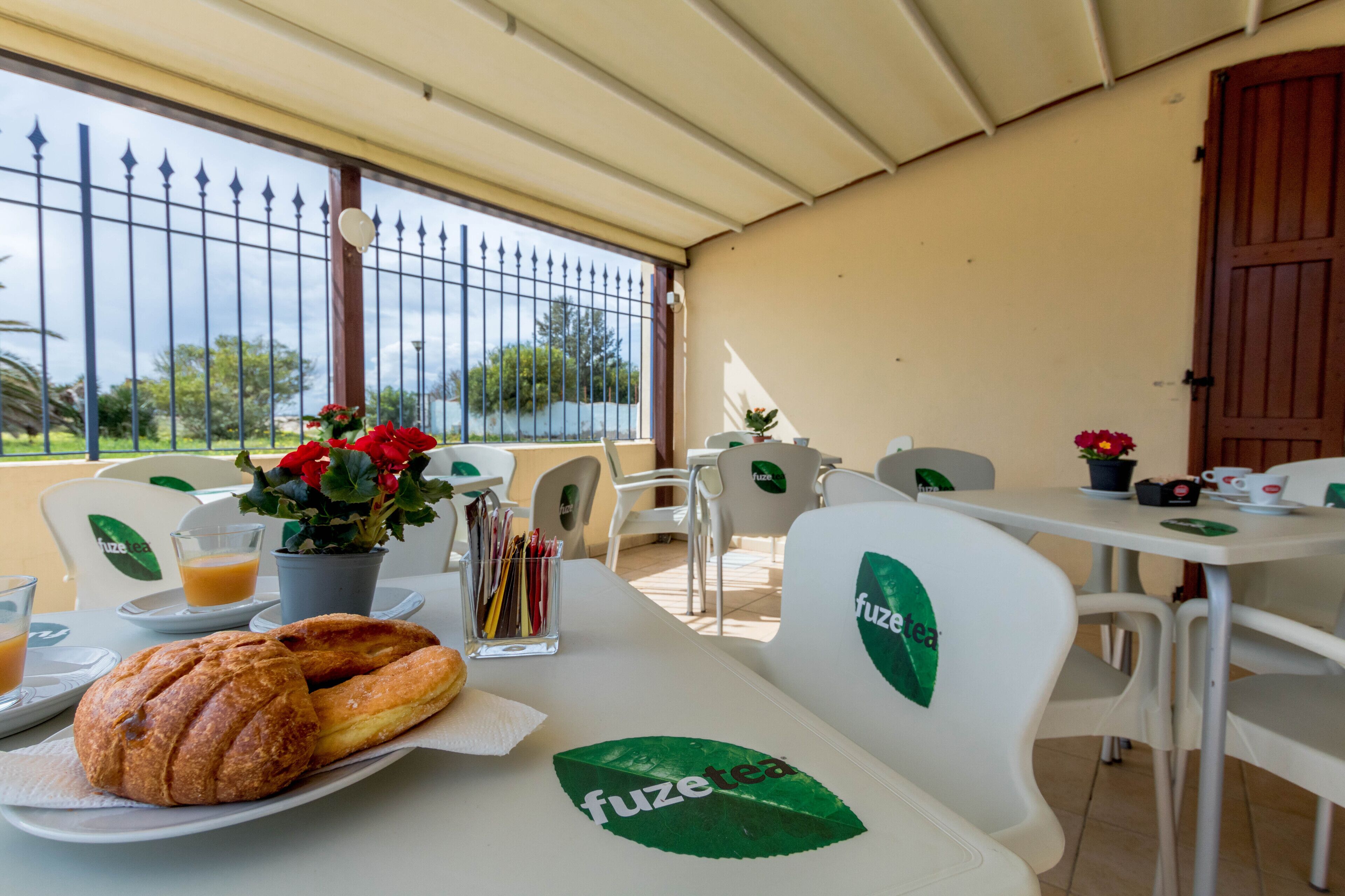 A table with a croissant and a glass of orange juice, chairs with 'fuzetea' branding, a potted plant with red flowers, and a view of the outdoors through a window with a metal fence.