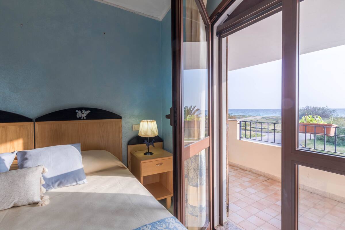 A bedroom with a bed, a nightstand, a lamp, and a view of the balcony and ocean.