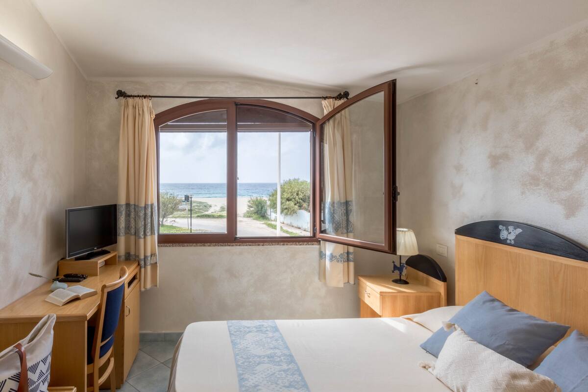 A hotel room with a bed, a desk with a TV, a window with curtains, and a view of the sea.