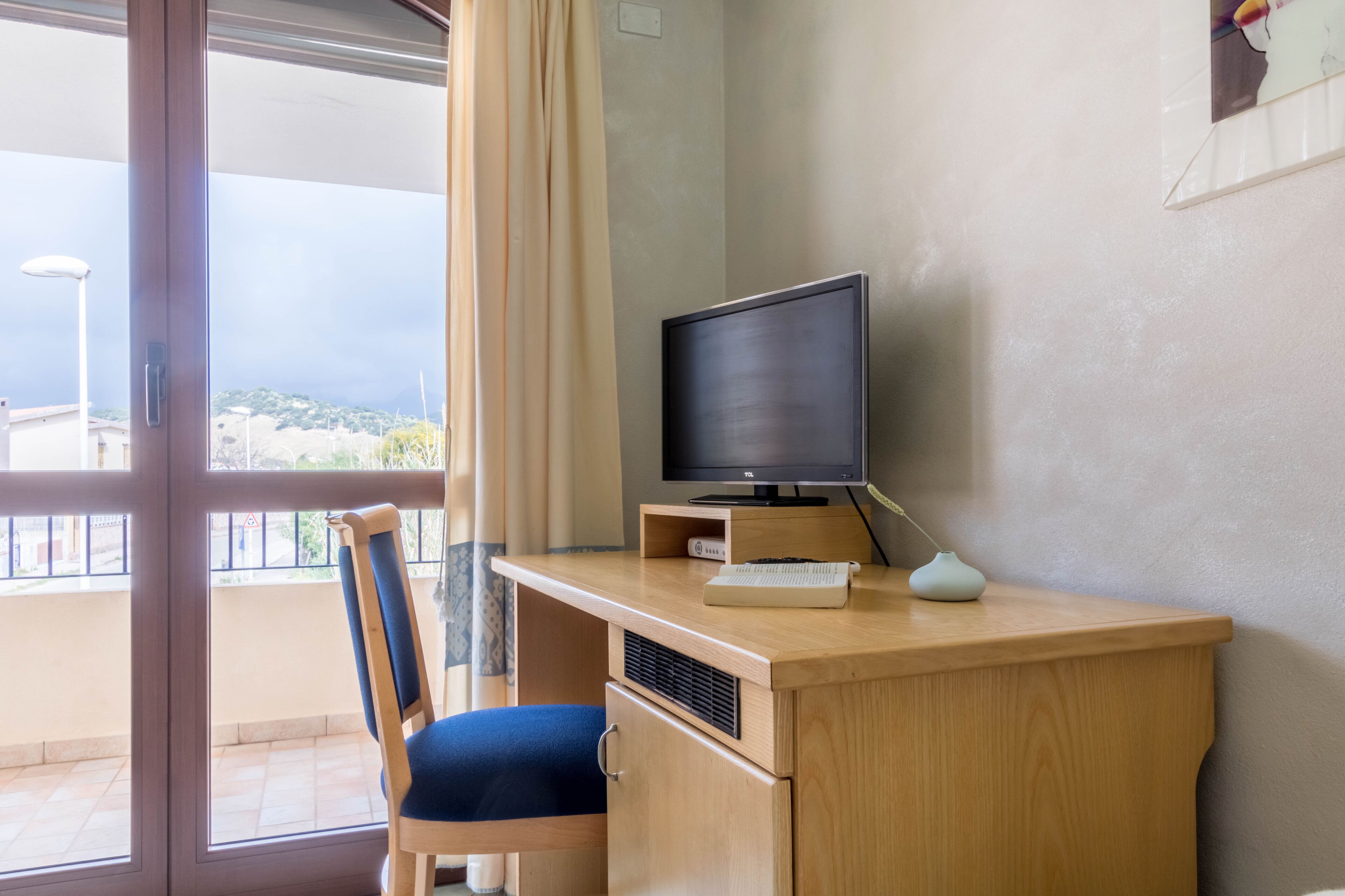 Een kamer met een houten bureau, een stoel, een televisie en een raam met uitzicht op gebouwen.