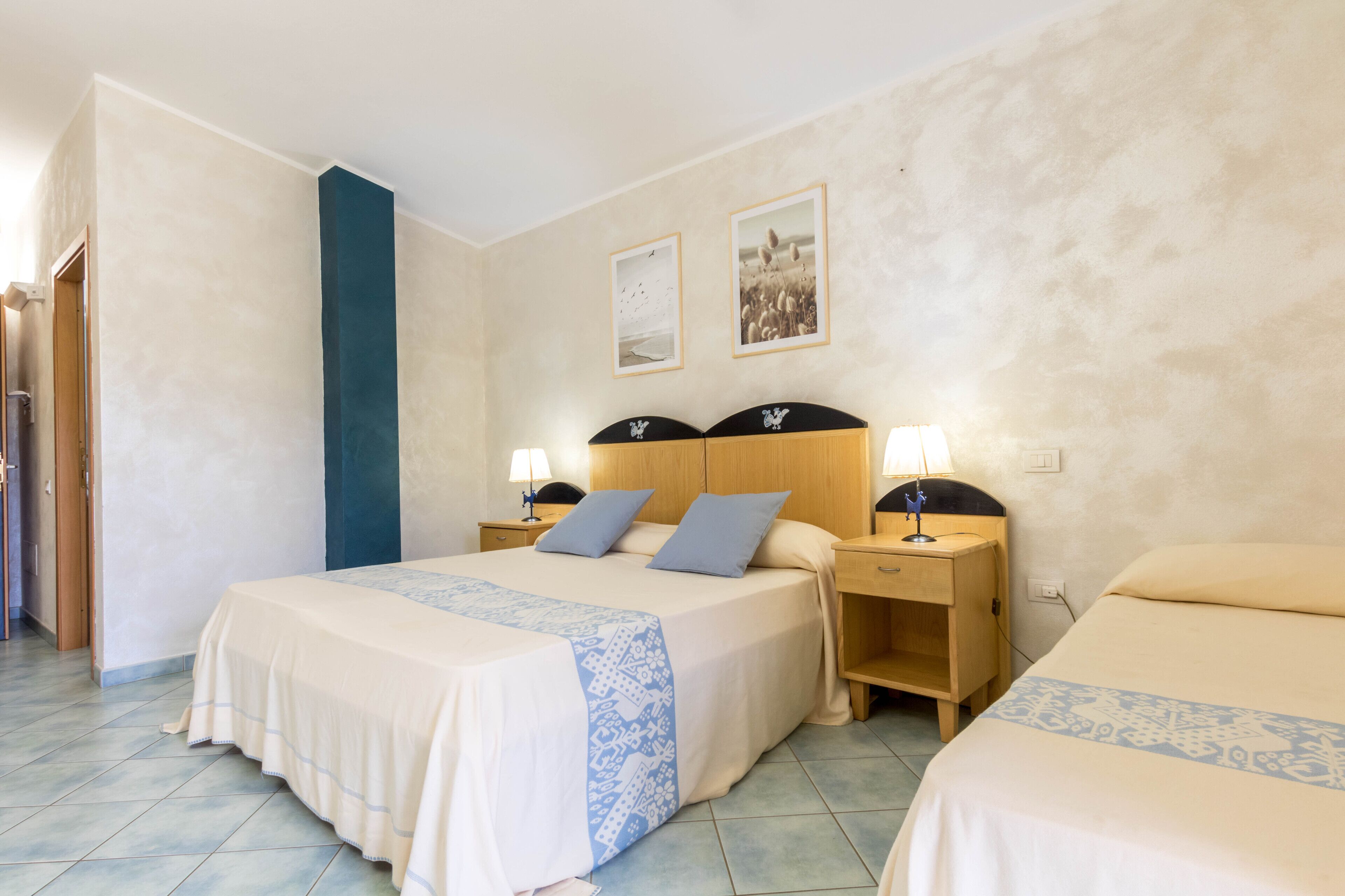 Quarto de hotel com duas camas, cabeceira de madeira, mesas de cabeceira com luminárias e uma porta à esquerda.