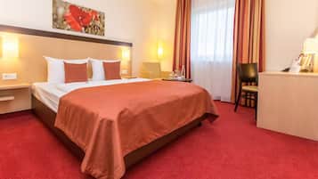 A hotel room with a large bed, a desk, and a chair. There is a painting on the wall and a window with curtains.