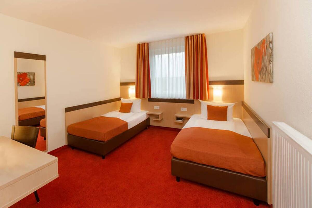 A hotel room with two single beds, a desk, a chair, and a large window with curtains.