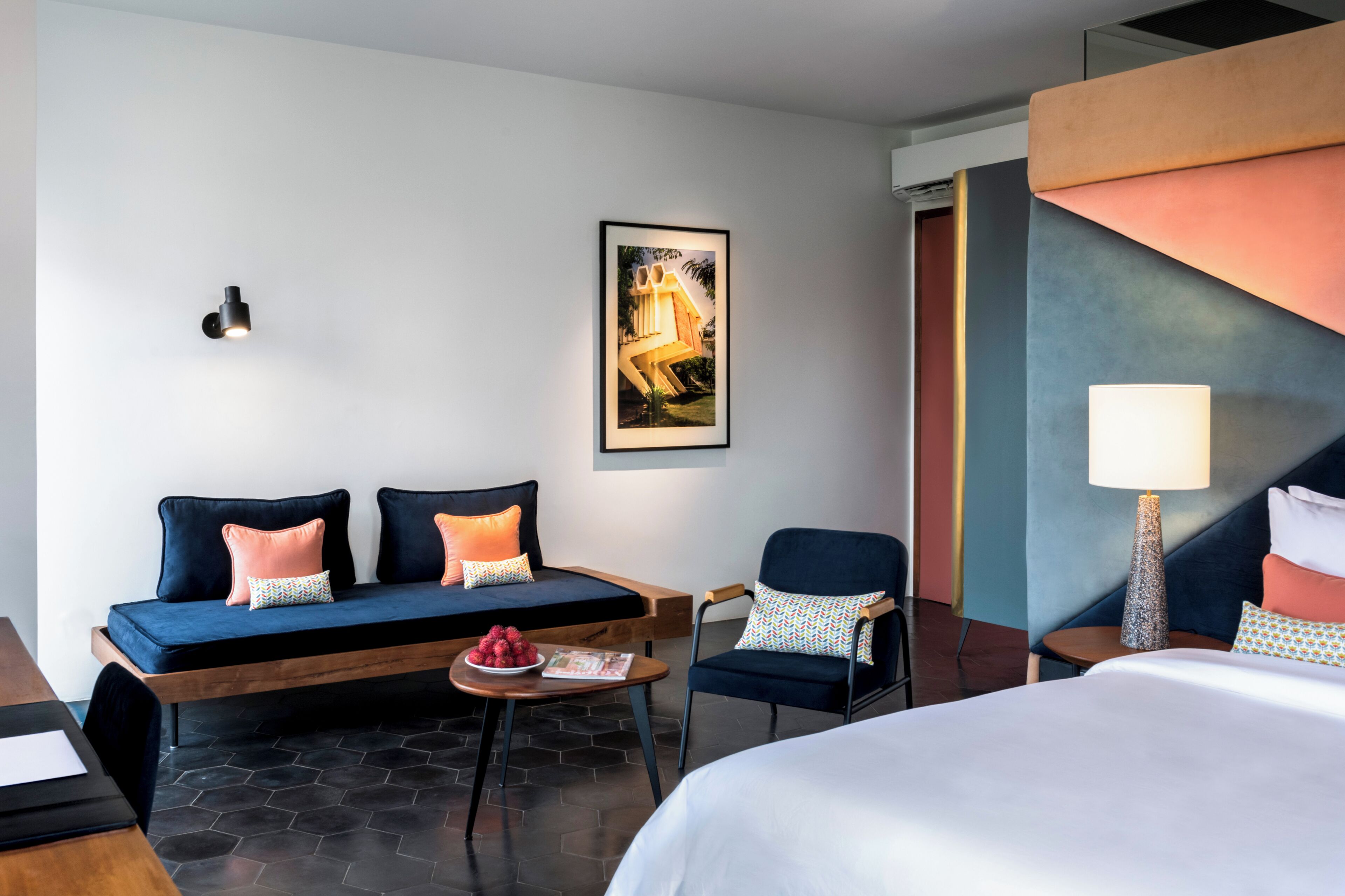 Un cuarto de hotel con una cama, un sofá, una silla, una mesita con un frutero, y un cuadro en la pared.