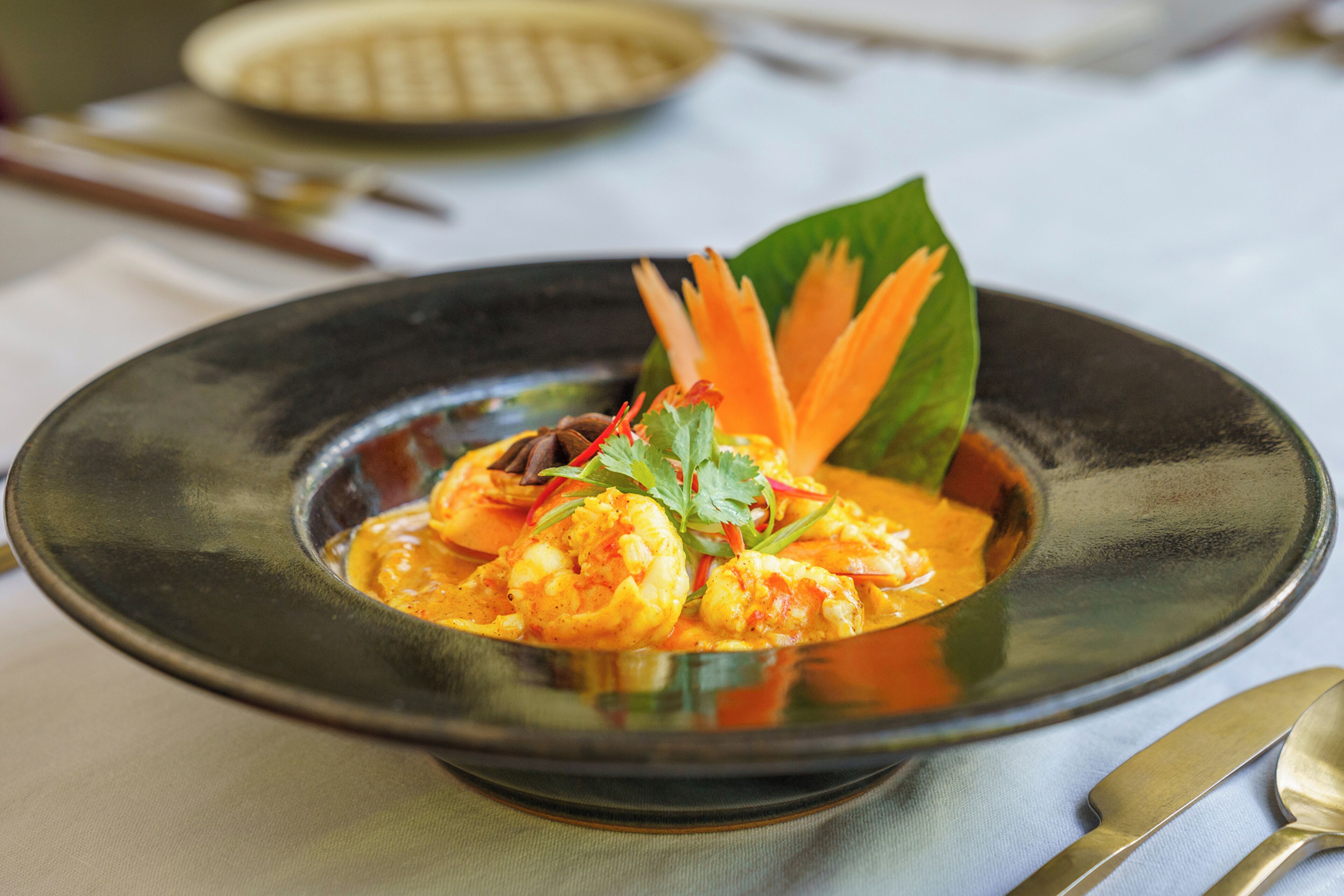 A bowl of shrimp curry with garnish, placed on a table with a napkin and cutlery.