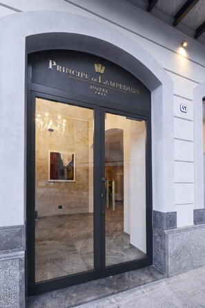 L’entrée de l’hôtel Prince de Lampedusa se distingue par une imposante porte en arc surmontée d’un lustre et dont la façade est recouverte d’une vitre réfléchissante.