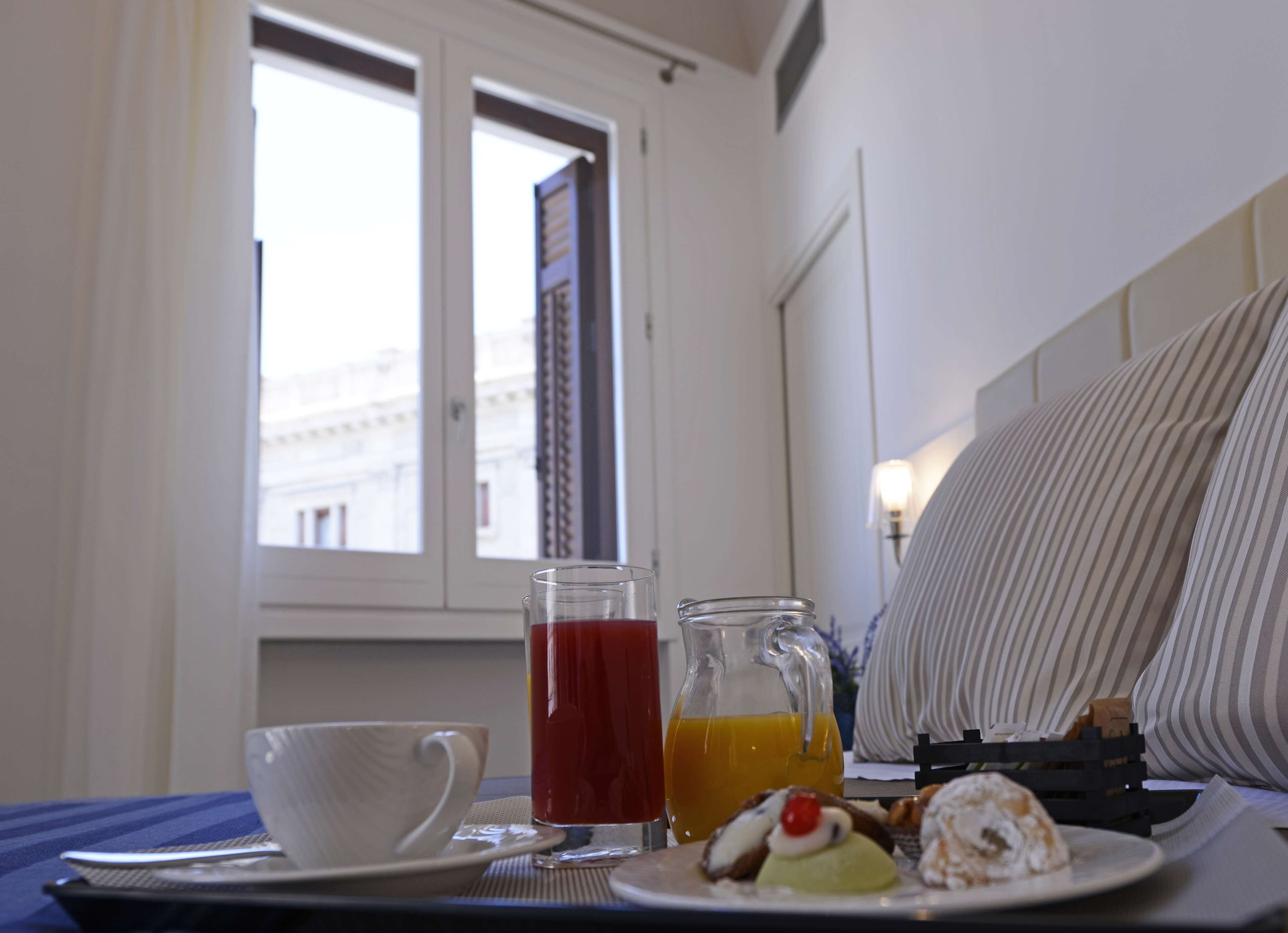 A breakfast tray with a cup, a glass of red juice, a glass of orange juice, and pastries on a table near a window with a view of buildings.