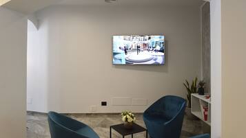 A lobby with a modern design, featuring a large flat-screen TV mounted on the wall, two blue armchairs, a small table with a vase of flowers, and a potted plant.