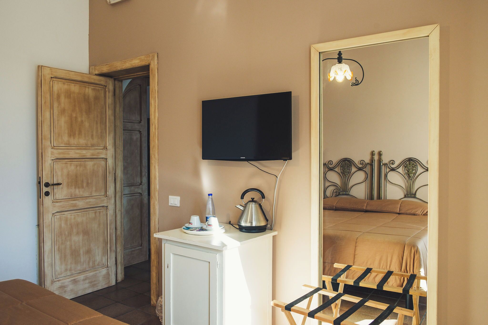Una habitación con puerta de madera, un televisor montado en la pared, una tetera sobre una mesita y una cama con cabecero.