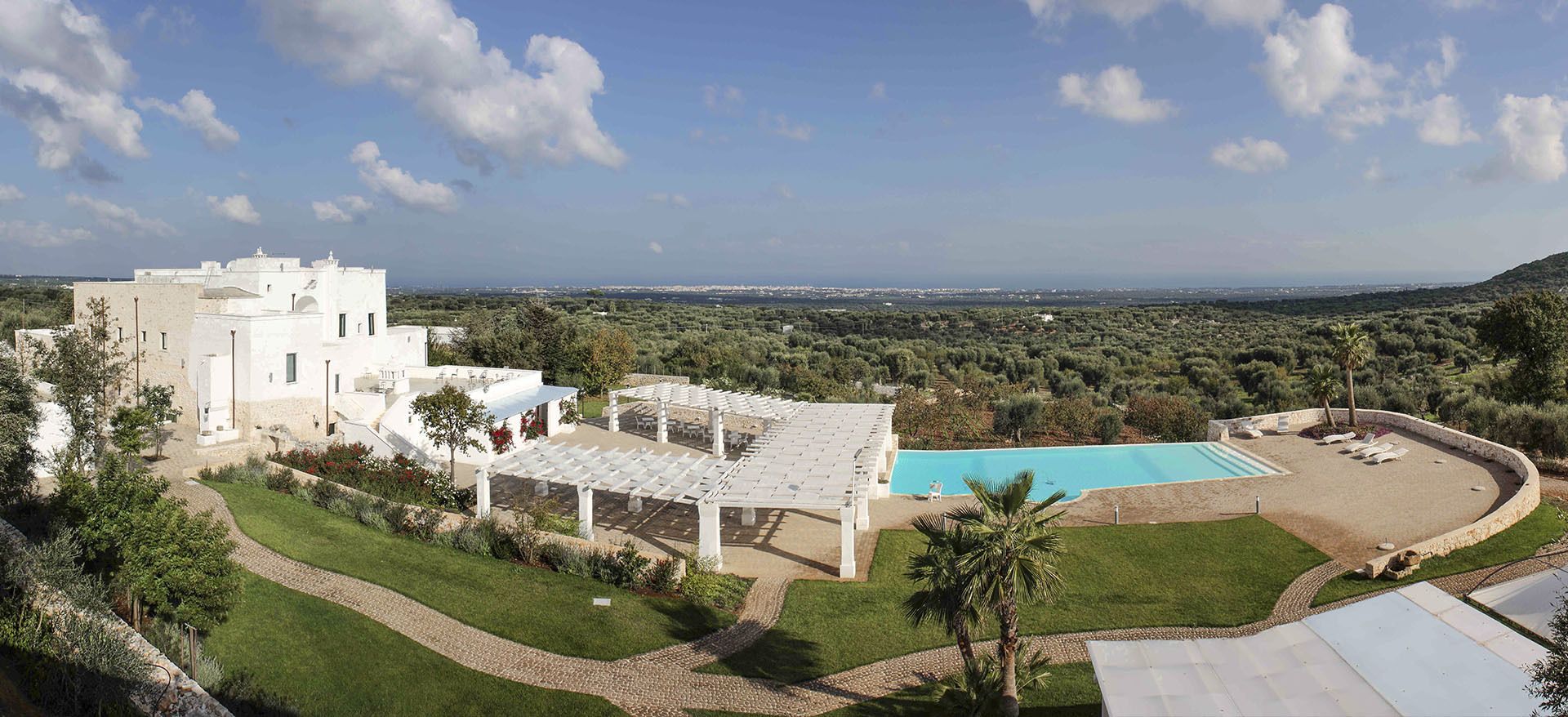 Un edificio grande y blanco con piscina y zona de estar al aire libre, rodeado de exuberante vegetación y vistas al campo.
