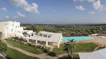 Un edificio grande y blanco con piscina y zona de estar al aire libre, rodeado de exuberante vegetación y vistas al campo.