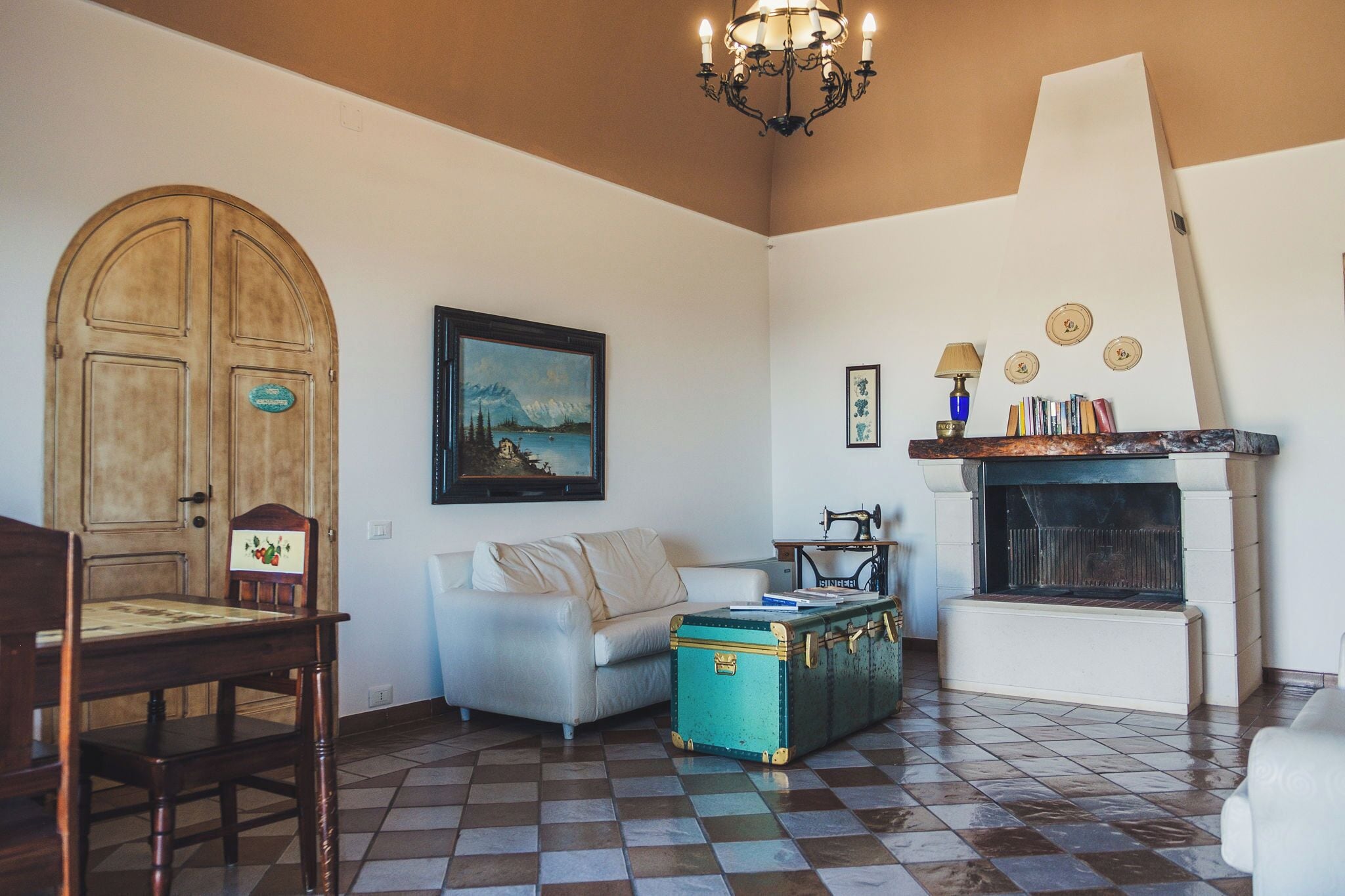 A room with a checkered floor, a wooden door, a white sofa, a green trunk, a fireplace, and a chandelier.
