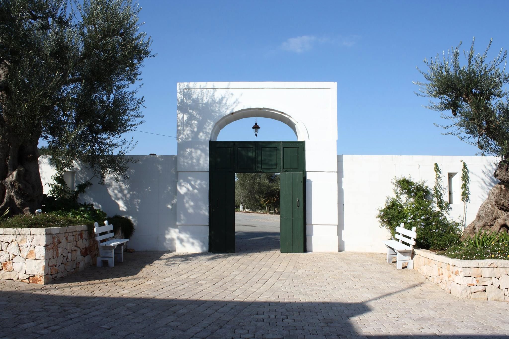A white building with a green door and an arched entrance. There are two white benches and a stone wall with greenery.